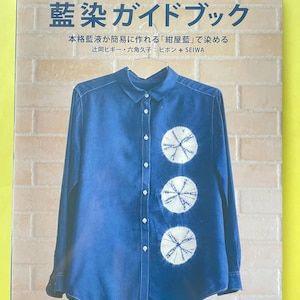 Könnte beinhalten: Ein Softcover-Buch mit dem Titel JAPAN BLUE und japanischem Text, das ein blaues langärmeliges Hemd mit Knopfleiste und drei weißen kreisförmigen Batikmustern auf der Vorderseite zeigt, das auf einem hellen Ziegelhintergrund hängt.