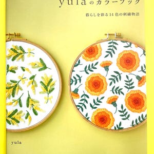 May include: A book titled yulaのカラーブック 暮らしを彩る14色の刺繍物語 with a light green cover. The cover features two circular wooden embroidery hoops containing white fabric with yellow mimosa and orange flower embroidery designs.