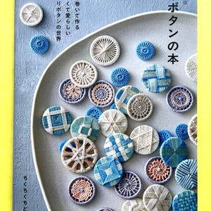 Könnte beinhalten: Ein Buchcover mit dem Titel 糸ボタンの本, das eine Sammlung runder, handgefertigter Garnknöpfe in verschiedenen Blau-, Beige- und Rosatönen zeigt. Die Knöpfe weisen komplexe geometrische Muster auf, die durch gewickeltes Garn erzeugt wurden.