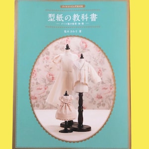 May include: A book titled "Model Paper Textbook" in Japanese, featuring illustrations of doll clothing and dress forms. The cover is teal with a gold border, and the background has a floral pattern.