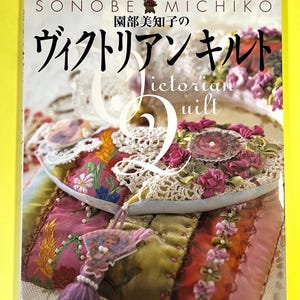 Puede incluir: Portada de libro con texto japonés y las palabras en inglés "Victorian Quilt". La portada presenta un primer plano de un edredón colorido y ornamentado con detalles en rosa y morado, encaje y adornos florales. El libro está sobre un fondo amarillo.