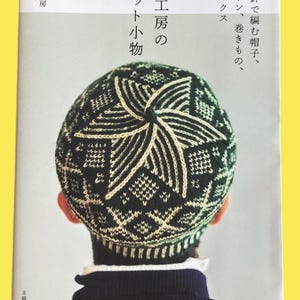 以下が含まれることがあります： 幾何学模様のダークグリーンとクリーム色のニット帽をかぶった人。帽子は「風工房のニット小物」というタイトルの日本の本の表紙に掲載されています。この本には、靴下、ミトン、帽子を編むことについてのテキストも含まれています。
