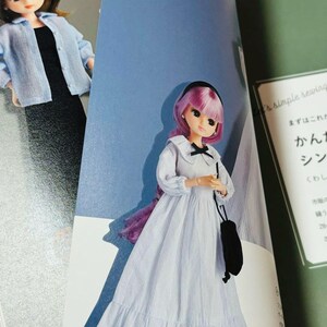Op de afbeelding: Een pop met roze haar en een zwarte haarband, gekleed in een wit en blauw gestreepte jurk met een zwarte strik en een zwarte handtas. De pop staat op een pagina van een boek met Japanse tekst.