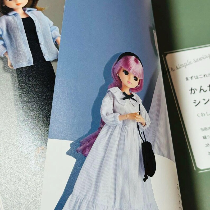 Op de afbeelding: Een pop met roze haar en een zwarte haarband, gekleed in een wit en blauw gestreepte jurk met een zwarte strik en een zwarte handtas. De pop staat op een pagina van een boek met Japanse tekst.