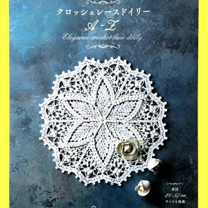 May include: A white crochet doily with a floral design, displayed on a dark blue background. The book cover reads "Elegance crochet lace doily A-Z" with Japanese text. The doily's diameter is 8.25 to 22.5 inches.