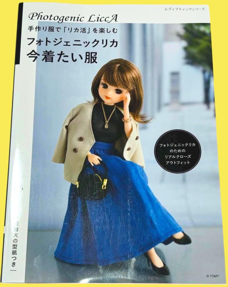 Op de afbeelding: Boekomslag met een pop in een zwarte top, blauwe rok en beige jasje. De boektitel is "Photogenic LiccA" in het Engels en Japans. De pop houdt een zwarte handtas vast en draagt zwarte schoenen.