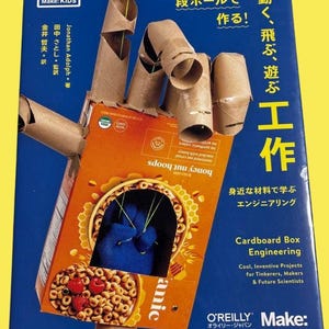 Könnte beinhalten: Ein Buch mit dem Titel "Cardboard Box Engineering" mit einem DIY-Kartonhandprojekt. Die Hand besteht aus Pappröhren und einer orangefarbenen Müslischachtel mit dem Text "honey nut hoops".