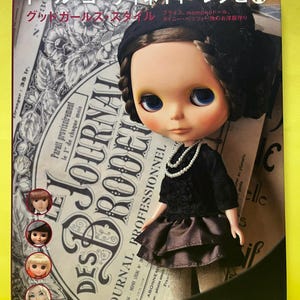 Op de afbeelding: Een Blythe-pop in een zwarte outfit met een parelketting, geplaatst voor een boekcover met Japanse tekst. Het boek heet "Dolly-Dolly Books". De achtergrond bevat vintage krant-achtige graphics.