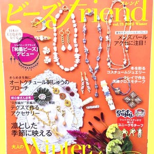 Może przedstawiać: Okładka magazynu prezentująca różne elementy biżuterii, w tym naszyjniki, kolczyki i broszki. Okładka zawiera tekst w języku japońskim i angielskim, z tytułem "Beads Friend" i rokiem 2022. Biżuteria jest prezentowana na żółto-pomarańczowym tle.