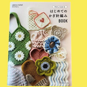 以下が含まれることがあります： 日本語のテキストと様々なかぎ針編みアイテムが掲載された本。かぎ針編みアイテムには、白いデイジーが付いた緑色のバッグ、ハート型の鍋つかみ、ミトン、花型のアイテムなどがあります。アイテムは様々な色です。