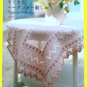 以下が含まれることがあります： ピンク色のハーダンガー刺繍のクローズアップ。花柄と幾何学模様が特徴で、ドイリーまたはテーブルランナーの可能性があります。縁はスカラップです。本には「ハーダンガー刺繍」と日本語で書かれています。背景には花瓶に入った花があります。