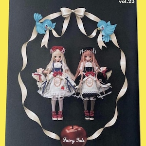 Op de afbeelding: De cover van het tijdschrift "Dollybird vol.23" toont twee poppen in gedetailleerde jurken, omlijst door een wit lint en blauwe vogelornamenten. Een rode appel met de tekst "Fairy Tale" staat onderaan. De achtergrond is zwart.