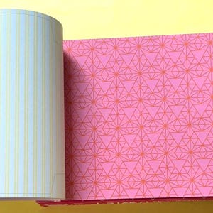 Puede incluir: Cuaderno abierto con p&aacute;ginas que muestran un patr&oacute;n geom&eacute;trico rosa y p&aacute;ginas rayadas. El patr&oacute;n rosa presenta un dise&ntilde;o repetitivo de tri&aacute;ngulos y l&iacute;neas en un tono m&aacute;s claro de rosa. Las p&aacute;ginas rayadas tienen l&iacute;neas verticales amarillas.