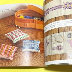 Puede incluir: Un libro abierto muestra varios proyectos de costura. Tres peque&ntilde;os cojines cuadrados con coloridos dise&ntilde;os de punto de cruz est&aacute;n dispuestos sobre una superficie de madera. Una caja de madera con suministros de costura est&aacute; en el fondo. Tambi&eacute;n se muestra un estuche para agujas.