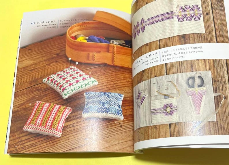 Puede incluir: Un libro abierto muestra varios proyectos de costura. Tres peque&ntilde;os cojines cuadrados con coloridos dise&ntilde;os de punto de cruz est&aacute;n dispuestos sobre una superficie de madera. Una caja de madera con suministros de costura est&aacute; en el fondo. Tambi&eacute;n se muestra un estuche para agujas.
