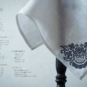 Op de afbeelding: Een wit linnen textiel met een donkerblauwe bloemen- en schedelborduurontwerp. Het textiel is gedrapeerd over een donkere houten tafel. De afbeelding toont ook tekst in het Japans.