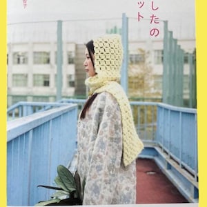 May include: A book cover featuring a woman wearing a light yellow knitted hood and scarf. The book title is in Japanese, with the author's name and publisher's logo visible. The background shows a blue railing and a building.