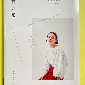 以下が含まれることがあります： 甘い服というタイトルの濱田明日香によるソフトカバー本。表紙には、黒い水玉模様の白いブラウスと赤いイヤリングを身につけた人物の写真が掲載されている。表紙には日本語のテキストと波線のイラストが描かれている。