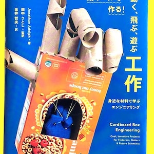 May include: A book cover titled "Cardboard Box Engineering" with a cardboard box hand construction. The cover is orange and blue, with text in English and Japanese. The hand is made of cardboard tubes and a cereal box.