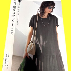 以下が含まれることがあります： ダークグレーのプリーツスカートのドレスを着た女性、サングラス、シルバーのネックレス。小さなクリーム色のハンドバッグを持っています。画像は日本語のテキストが入った日本のファッション雑誌からのものです。