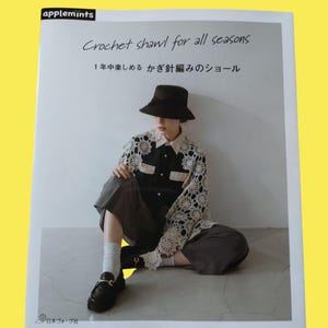 May include: A book cover with the text "Crochet shawl for all seasons" and Japanese text. The image shows a person wearing a black shirt with a cream-colored crochet shawl, dark pants, and a black hat. The person is sitting down.