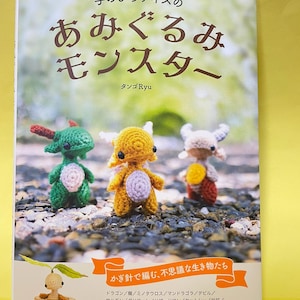 以下が含まれることがあります： 「手のひらサイズのあみぐるみモンスター」というタイトルの書籍の表紙。緑、黄色、ベージュの3体の小さな編みぐるみのモンスターが写っており、左下にはマンドラゴラの編みぐるみがある。表紙には日本語のタイトルと、掲載されているクリーチャーの名前のリストが記載されている。