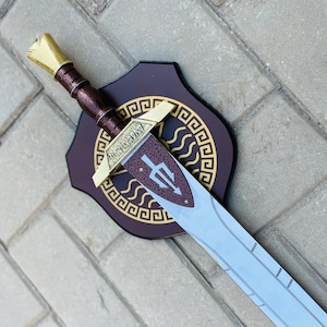 May include: A decorative sword with a gold hilt and a silver blade, mounted on a dark brown shield with gold accents. The sword features a brown handle and a gold crossguard. The shield has a decorative pattern.