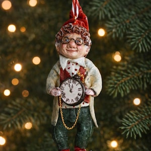 Könnte beinhalten: Ein skurriler Weihnachtsschmuck, der eine lächelnde Figur mit lockigem braunem Haar, runder Brille und roter Mütze darstellt. Die Figur trägt einen cremefarbenen Mantel, eine Weste mit Herzmustern und eine grüne Hose. Sie hält eine Taschenuhr und rot-weiß gestreifte Schuhe.