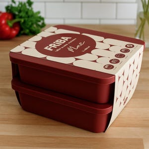 Può includere: Un contenitore per alimenti bordeaux a due livelli con una custodia decorativa in cartone. La custodia presenta il testo "FRIBA" e "More" in un design circolare. Il contenitore è su una superficie di legno.