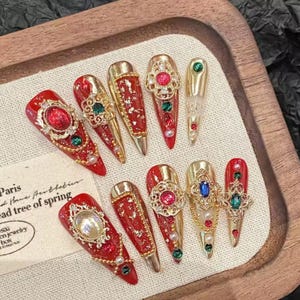 Peut inclure: Collection de faux ongles longs et ornés, rouges et dorés. Chaque ongle est décoré de dorures, de bijoux et de perles. Certains ongles ont une base transparente avec des accents dorés. Les ongles sont présentés sur un plateau en bois.