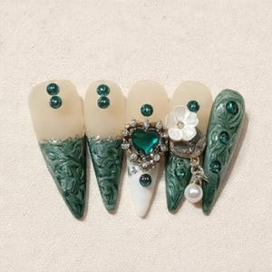 Peut inclure: Ensemble de cinq faux ongles longs en forme d'amande. Les ongles présentent divers motifs, notamment des paillettes vertes et argentées, des pierres précieuses vert émeraude, un bijou en forme de cœur, une fleur blanche et un charme de perle pendante.