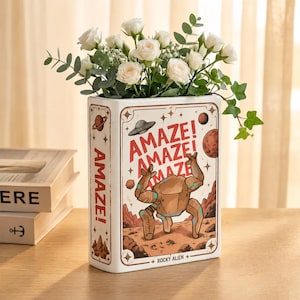 Pode incluir: Um vaso de cerâmica retangular em forma de livro, apresentando um gráfico de uma criatura alienígena castanha numa paisagem rochosa com o texto AMAZE! e ROCKY ALIEN. O vaso está cheio de rosas brancas e folhas de eucalipto.