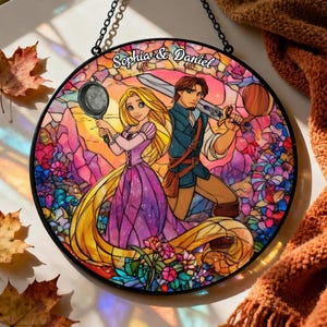 Op de afbeelding: Een rond gebrandschilderd glazen kunstwerk met een prinses en een prins die een koekenpan en een zwaard vasthouden. Het kunstwerk is kleurrijk, met een bloemenachtergrond en de tekst "Sophia & Daniel" bovenaan.