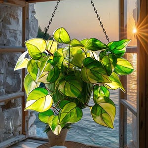 May include: A stained glass hanging planter with a vibrant green and yellow pothos plant. The leaves are detailed with white accents and are suspended by a black chain. The planter is set against a window with a view of the ocean.