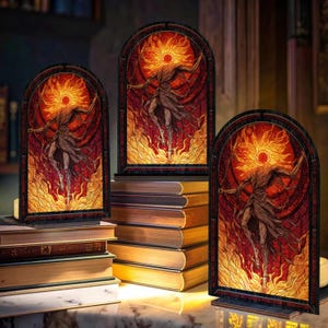 May include: Three arched stained-glass style panels depicting a figure with outstretched arms, surrounded by flames and a sunburst. The panels are set on wooden bases and rest on stacks of books. The artwork features red, orange, and brown hues.