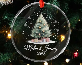 May include: A clear, round Christmas ornament with a red ribbon. The ornament features a watercolor-style Christmas tree with gold ornaments, gifts, and the text "Mike & Jenny 2025". Snowflakes are scattered around the tree.