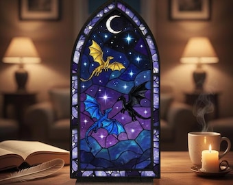 May include: A gothic arch stained glass window depicting a night sky with a crescent moon, stars, and three dragons in gold, blue, and black. A candle and a cup of coffee sit on a wooden table. The window has a dark frame.