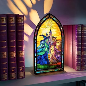 May include: A stained glass window depicting two figures, one in a witch's hat and the other in a crown, with a castle in the background. The window is set in a dark frame and sits on a shelf next to purple books.