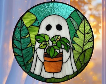 May include: Round stained glass window art featuring a white ghost holding a potted plant. The ghost has two black eyes and is surrounded by green leaves and a green border. The plant pot is orange.