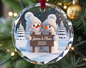 May include: A clear, faceted glass Christmas ornament featuring two snowmen wearing white knit hats and holding a wooden sign that reads "James & Olivia 2025". The ornament hangs from a red ribbon and is set against a snowy winter scene.