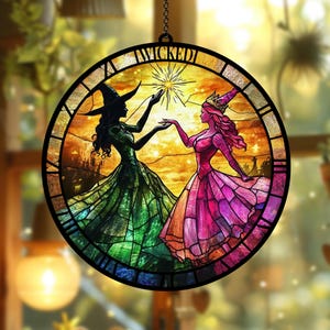Puede incluir: Vidriera redonda con dos siluetas, una con sombrero de bruja y vestido verde, la otra con vestido rosa y corona. La palabra "WICKED" está en la parte superior, con una estrella en el centro.