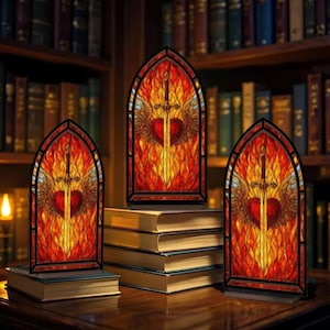 Op de afbeelding: Drie glas-in-loodpanelen met een hart doorboord door een zwaard, vleugels en vlammen. De panelen hebben een gotische boogvorm met een zwarte rand. De panelen staan op boeken voor een boekenkast.