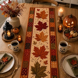 Peut inclure: Un chemin de table matelassé rectangulaire présentant un panneau central avec des feuilles d'érable appliquées dans des tons de rouge, orange et marron, bordé par un motif en patchwork de petits carrés aux couleurs automnales, incluant des motifs orange, verts et marron.