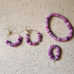 May include: A jewelry set featuring a bracelet, two pairs of hoop earrings, and a ring. The jewelry is made of small, flat, round beads in shades of purple and pink. The earrings and ring have gold-colored metal findings.