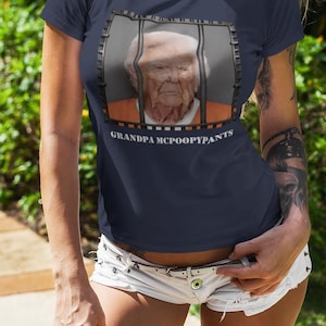 May include: Navy blue t-shirt featuring a graphic of an older man behind bars with the text "GRANDPA MCPOOPYPANTS". The shirt is short-sleeved and is being worn outdoors.