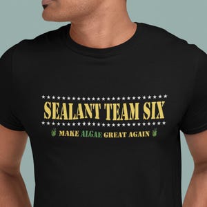 May include: A black short-sleeved t-shirt featuring the yellow text SEALANT TEAM SIX centered on the chest, flanked by rows of white stars. Below this, the text MAKE ALGAE GREAT AGAIN is printed in yellow, accompanied by small green icons.