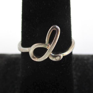 May include: A silver-toned ring featuring a cursive letter L design. The band is thin and metallic, with the script letter forming the centerpiece of the ring. The metal has a smooth, polished finish that reflects light across its curved surface.