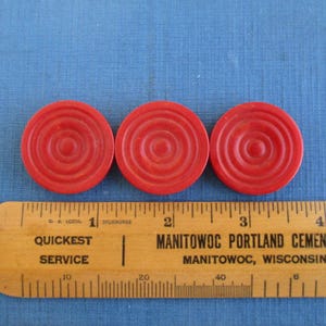 May include: Three red plastic game chips with concentric circles. The chips are about 1 inch in diameter and are lined up on a wooden ruler.