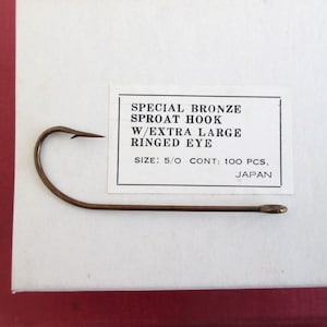 May include: A bronze Sproat hook with an extra-large ringed eye, size 5/0, is displayed on a white surface. The text on the label reads "SPECIAL BRONZE SPROAT HOOK W/EXTRA LARGE RINGED EYE SIZE: 5/0 CONT: 100 PCS. JAPAN".