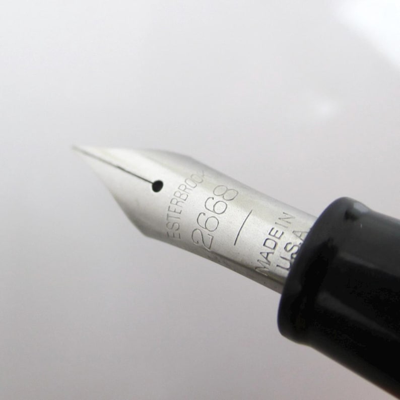 May include: Close-up of a silver fountain pen nib with the text "ESTERBROOK 2668 MADE IN U.S.A." The nib is sharp and pointed, with a small hole near the tip. The pen's body is black.